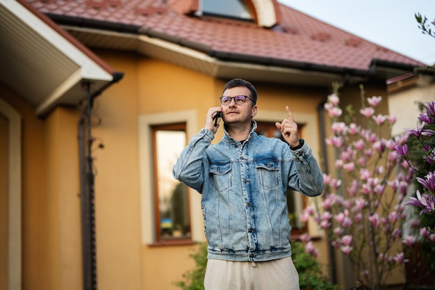 Portret stylowego mężczyzny w okularach i dżinsowej kurtce z telefonem komórkowym postawionym przed jego domem i pokazującym palec w górę