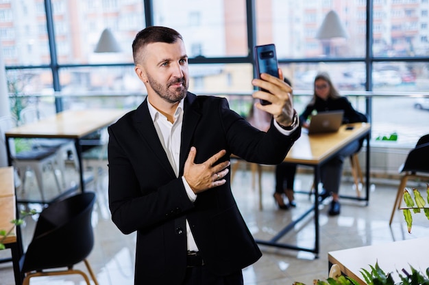 Portret stylowego młodego biznesmena stojącego w dużym nowoczesnym biurze na najwyższym piętrze i patrzącego na telefon