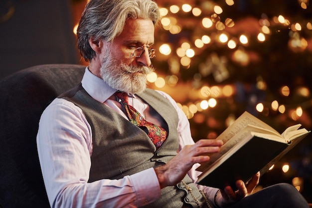 Portret stylowego seniora z siwymi włosami i brodą czytającą książkę w urządzonym pokoju bożonarodzeniowym.