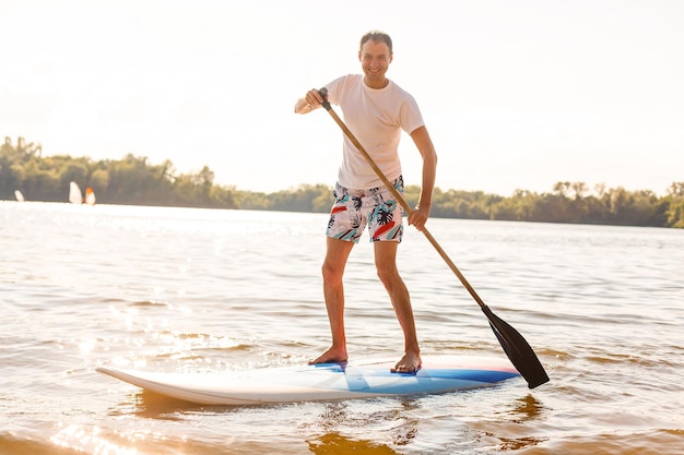 Portret surfera z deską SUP na plaży. Młody człowiek na paddleboard o świcie. Pojęcie sportów ekstremalnych. Styl życia męskiego surfera.