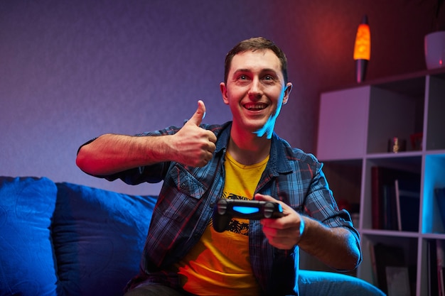 Portret szalonego, figlarnego gracza, chłopca grającego w gry wideo w pomieszczeniu, siedzącego na sofie, trzymając w rękach kontroler konsoli. Odpoczynek w domu, miłego weekendu