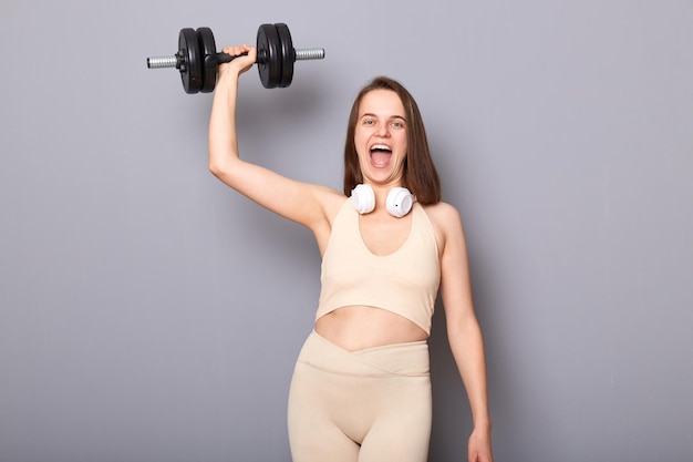 Portret szalonej podekscytowanej silnej kobiety ubranej w top i legginsy trzymającej w ręku sztangę podniosła rękę, wykonując ćwiczenia na triceps i biceps na szarym tle