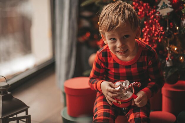 Portret szczere szczęśliwe dziecko w czerwonej piżamie w kratę trzymaj świąteczny kubek z piankami i trzciną cukrową
