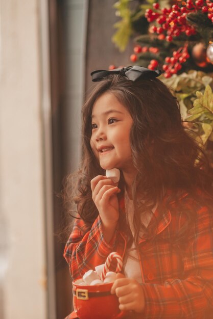 Portret szczerej azjatyckiej uśmiechniętej dziewczynki trzyma czerwony świąteczny kubek z piankami i trzciną cukrową