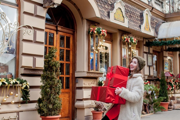 Portret Szczęśliwa, dobrze wyglądająca kobieta trzyma w dłoniach pudełko z prezentami i uśmiecha się, stojąc na zewnątrz Piękna szczęśliwa dziewczyna pozuje z prezentami świątecznymi Przygotowania do Nowego Roku