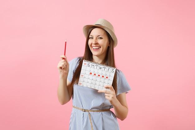 Portret szczęśliwa kobieta w niebieskiej sukience, kapeluszu trzymającym czerwony ołówek, kalendarz kobiet okresów do sprawdzania dni menstruacji na białym tle na różowym tle. Opieki zdrowotnej, koncepcja ginekologiczna. Skopiuj miejsce