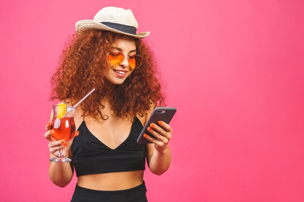 Portret szczęśliwa kobieta w okularach przeciwsłonecznych za pomocą smartfona i trzymając koktajl na białym tle