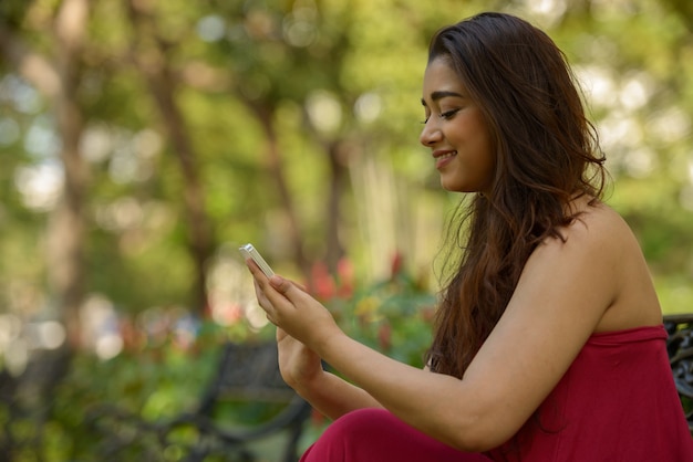 Portret szczęśliwa młoda piękna indyjska kobieta przy użyciu telefonu w parku