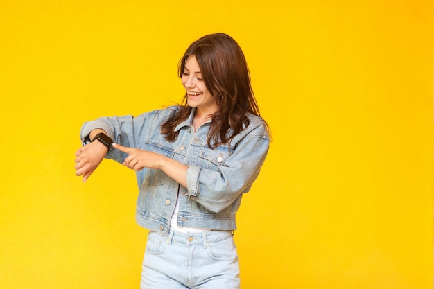 Portret szczęśliwa piękna brunetka młoda kobieta w denim stylu casual, stojąca, uśmiechnięta ząb, dotykająca i sprawdzająca jej inteligentny gadżet. kryty strzał studio, na białym tle na żółtym tle.