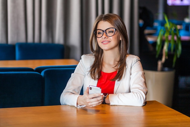 Portret szczęśliwa piękna stylowa brunetka młoda kobieta w okularach siedzi z inteligentny telefon komórkowy i odwracając z uśmiechem toothy i marzy kryty strzał studio kawiarnia tło biurowe