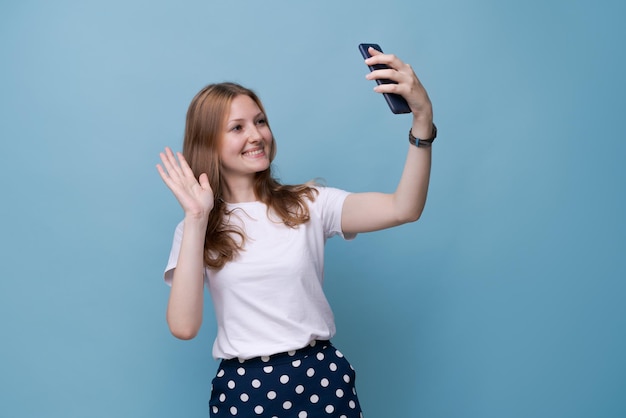 Portret szczęśliwa przyjazna młoda kaukaska dziewczyna w białej koszulce na niebieskim tle robi selfie i wa...