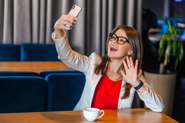 Portret szczęśliwa radosna piękna stylowa brunetka młoda kobieta w okularach siedzi na wideorozmowie lub robi selfie z toothy uśmiechem i szczęściem macha ręką kryty studio strzał kawiarnia biuro tło