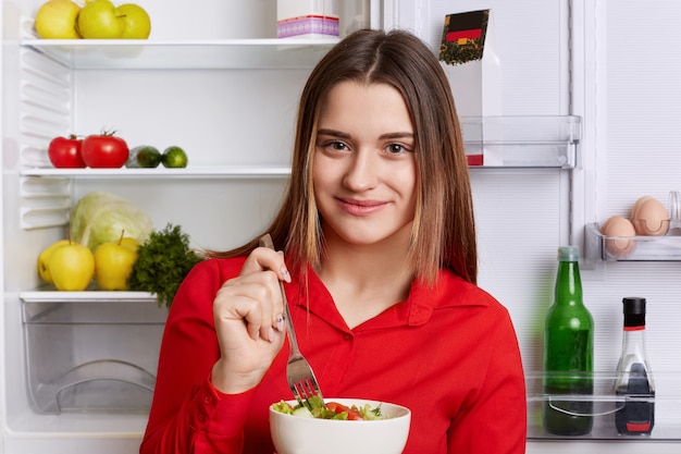 Portret szczęśliwa uradowana młoda kobieta o atrakcyjnym wyglądzie ubrana w czerwoną koszulę, je pyszną sałatkę warzywną, stoi w kuchni przy otwartej lodówce, trzyma się diety. Koncepcja zdrowego żywienia