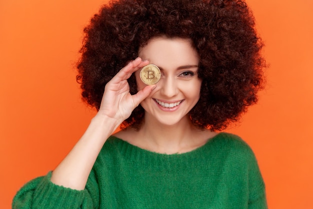 Portret szczęśliwa uśmiechnięta kobieta z fryzurą afro na sobie zielony sweter w stylu casual zakrywający oko ze złotym bitcoinem koncepcja blockchain kryty strzał studio na białym tle na pomarańczowym tle