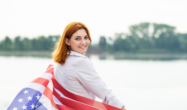 Portret szczęśliwa uśmiechnięta rudowłosa dziewczyna z flagą narodową USA na ramionach. Młoda kobieta obchodzi Dzień Niepodległości Stanów Zjednoczonych.