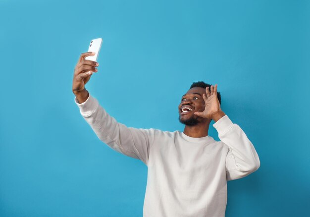 Portret szczęśliwego Afroamerykanina robiącego selfie i uśmiechającego się na niebieskim, odizolowanym tle