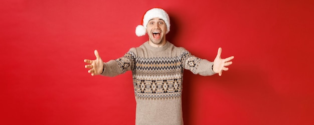 Portret szczęśliwego, atrakcyjnego mężczyzny w świątecznym swetrze i santa hat, sięgającego po prezent, chce coś wziąć i uśmiechniętego, stojącego na czerwonym tle