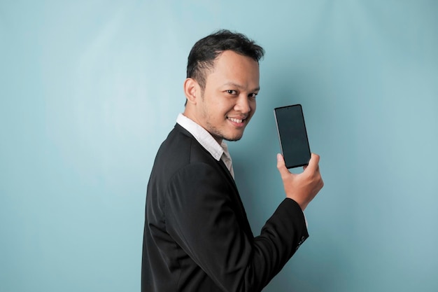 Portret szczęśliwego azjatyckiego biznesmena uśmiecha się i trzyma smartfona pokazując miejsce na kopię na ekranie, ubrany w czarny garnitur na białym tle na niebieskim tle