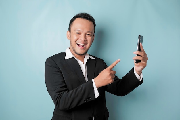 Portret szczęśliwego azjatyckiego biznesmena uśmiecha się i trzyma swojego smartfona w czarnym garniturze odizolowanym niebieskim tłem