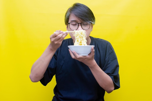 Portret szczęśliwego azjatyckiego mężczyzny w czarnej koszulce zjada makaron instant za pomocą pałeczek i izolowanej miski