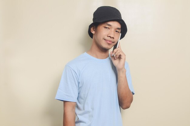 Portret szczęśliwego azjatyckiego młodego człowieka noszącego czarny kapelusz, podczas gdy na telefon na białym tle