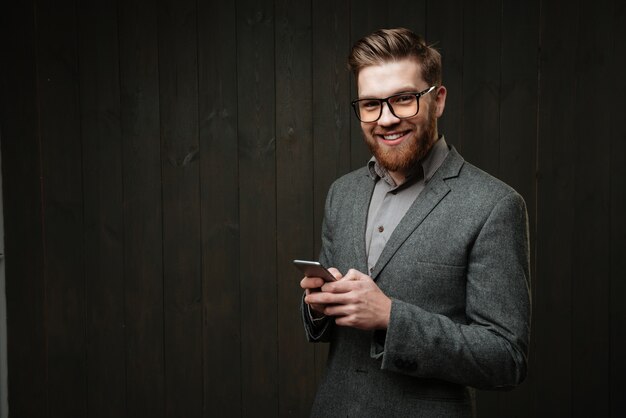 Portret szczęśliwego brodatego mężczyzny w garniturze na co dzień, trzymającego telefon komórkowy