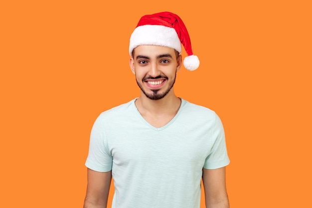 Portret szczęśliwego brodatego młodzieńca w kapeluszu świętego mikołaja i dorywczo białej koszulce stojącej, patrząc na kamerę z uśmiechem zębów, ciesząc się świątecznymi. studio strzał na białym tle na pomarańczowym tle