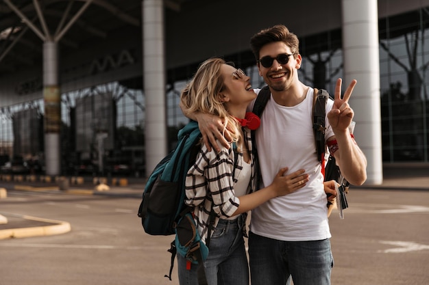 Portret Szczęśliwego Brunetki W Okularach Przeciwsłonecznych Pokazujących Znak Pokoju I Stylowej Blondynki W Pobliżu Lotniska Urocza Kędzierzawa Dziewczyna Przytula Chłopaka I Trzyma Plecak