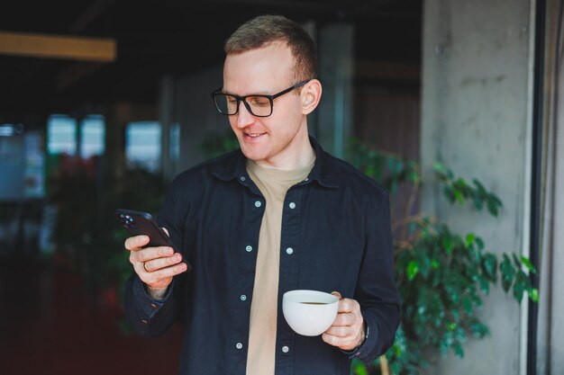 Portret Szczęśliwego Człowieka Sukcesu W Okularach Optycznych, Trzymającego W Rękach Nowoczesny Telefon Komórkowy I Kawę, Wesoły Bloger Z Cyfrowym Smartfonem Pozującym W Kawiarni