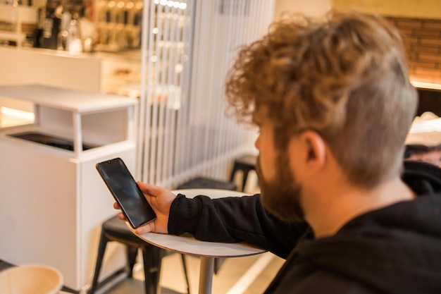 Portret szczęśliwego faceta, który dokonuje transakcji pieniężnych za pośrednictwem aplikacji na telefon komórkowy z internetem 4g, siedząc w kawiarni wieczorem na ulicy, poważny tysiącletni mężczyzna czytający wiadomość tekstową na smartfonie