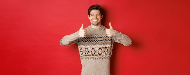 Portret szczęśliwego i zadowolonego przystojnego faceta w bożonarodzeniowym swetrze pokazującym kciuk do góry i kiwającym głową z aprobatą uśmiechnięty zadowolony stojąc na czerwonym tle