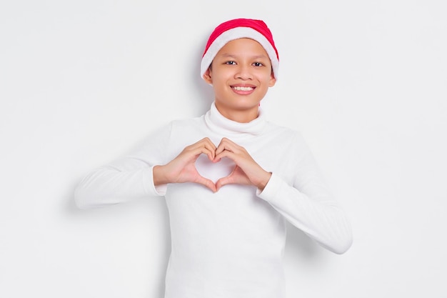Portret szczęśliwego małego azjatyckiego człowieka w świątecznym kapeluszu pokazującym gest serca z dwiema rękami na białym tle nad białym tłem Szczęśliwego nowego roku 2023 koncepcja wakacje