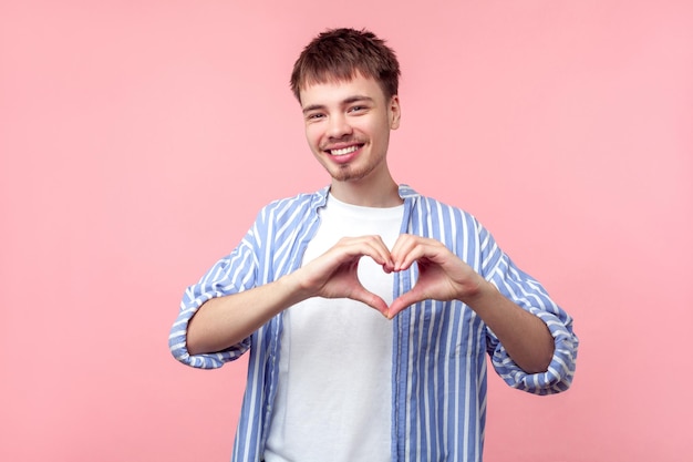 Portret szczęśliwego miłosnego brązowowłosego mężczyzny z małą brodą i wąsami w casualowej koszuli w paski uśmiechający się i robiący serce z rękami symbol miłości studio strzał na białym tle na różowym tle