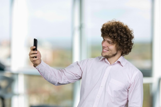 Zdjęcie portret szczęśliwego młodego mężczyzny z kręconymi włosami robiącego selfie za pomocą smartfona w oknie wewnętrznym