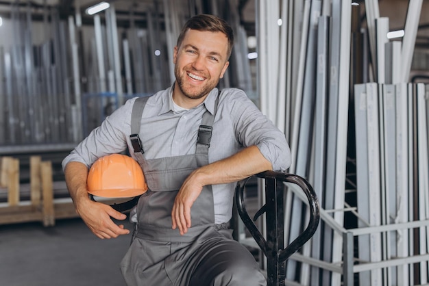 Zdjęcie portret szczęśliwego pracownika w pomarańczowym kasku i kombinezonie trzymającego hydrauliczną ciężarówkę na tle fabryki i aluminiowych ram