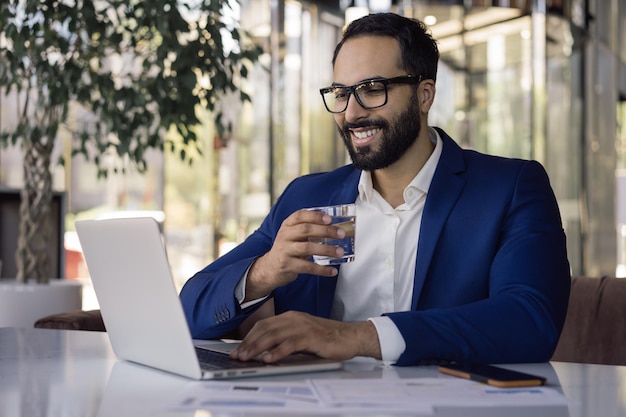 Portret szczęśliwego przystojnego menedżera korzystającego z wody pitnej laptopa siedzącego w biurze