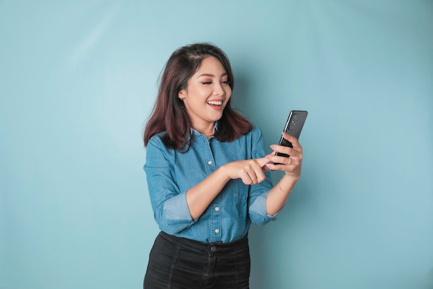 Portret szczęśliwej Azjatki uśmiecha się i trzyma swojego smartfona w niebieskiej koszuli odizolowanej na niebieskim tle