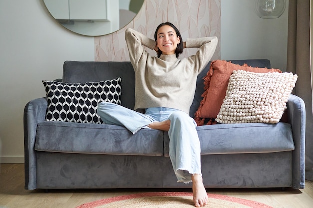 Portret szczęśliwej azjatyckiej kobiety, która czuje się leniwa, rozciągając się na kanapie i uśmiechając się, zadowolona relaksując się w domu r
