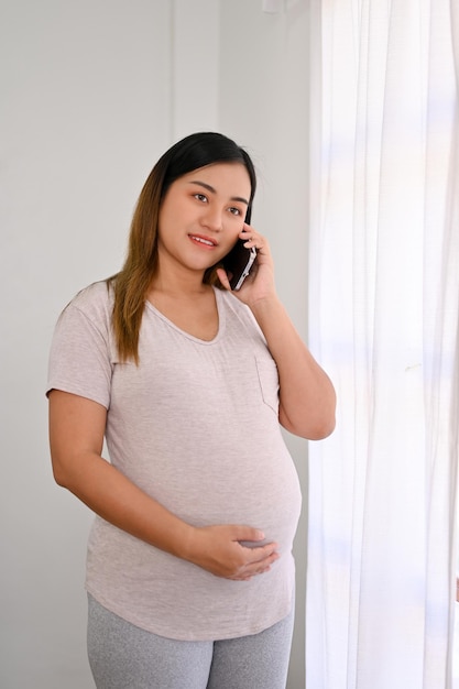 Portret szczęśliwej azjatyckiej kobiety w ciąży stoi przy oknie przez telefon z mężem