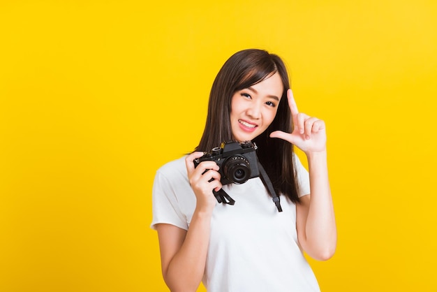 Portret szczęśliwej azjatyckiej pięknej młodej kobiety fotograf trzymającej na rękach cyfrowy aparat fotograficzny bez lusterka, studio strzał odizolowane na żółtym tle, styl życia nastolatka hobby koncepcja podróży