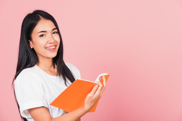 Portret szczęśliwej azjatyckiej pięknej młodej kobiety pewnej siebie uśmiechniętej stojącej trzymającej otwartą pomarańczową książkę lub pamiętnik do czytania, studio strzał odizolowane na różowym tle, z miejsca kopiowania, koncepcja edukacji
