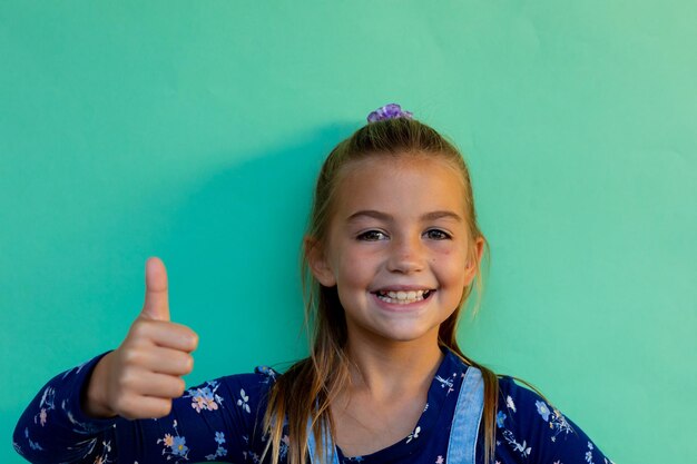 Zdjęcie portret szczęśliwej białej uczennicy z kciukiem w górze na niebieskim tle w szkole podstawowej