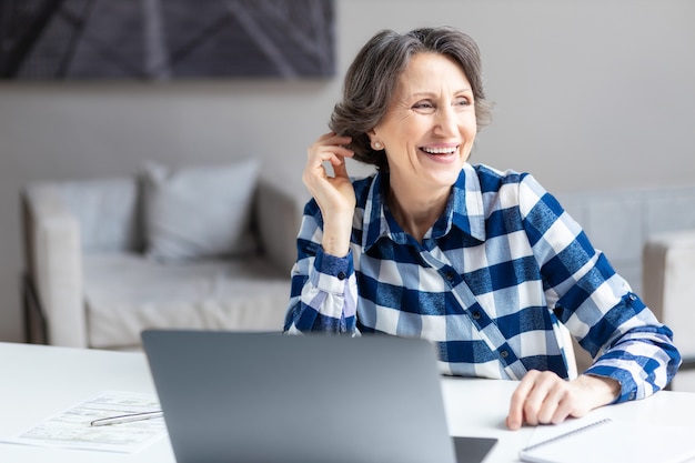 Portret szczęśliwej biznesowej w wieku kobiety z laptopem siedzącej przy biurku w biurze lub w domu odwraca wzrok od okna i uśmiecha się