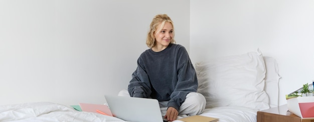 Portret szczęśliwej blondynki pracującej w domu, siedzącej na łóżku z laptopem i notatnikami