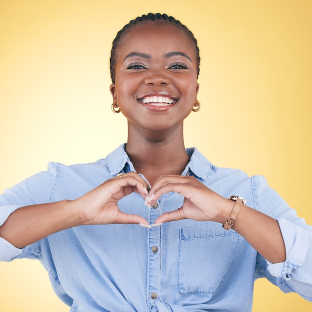 Zdjęcie portret szczęśliwej czarnej kobiety i dłonie z sercem do opieki miłosnej lub wsparcia na żółtym tle studia twarz afrykańskiej kobiety uśmiecha się z kochającym kształtem emoji lub ikoną symbolu na walentynki