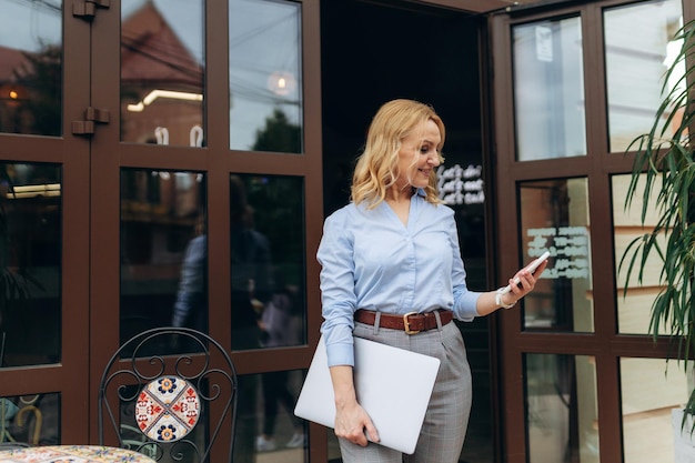 Portret szczęśliwej dojrzałej bizneswoman korzystającej z telefonu stojąc na chodniku kawiarni
