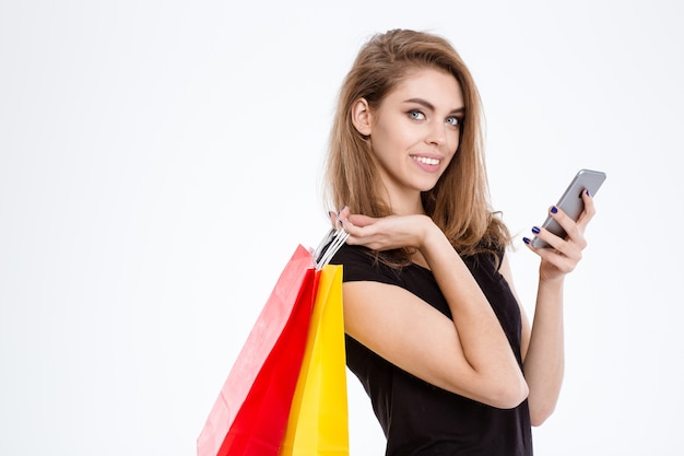Portret szczęśliwej kobiety trzymającej torby na zakupy i używającej smartfona na białym tle