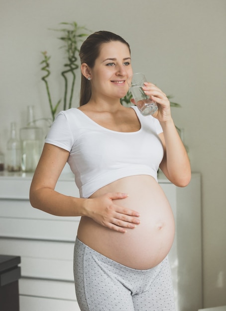 Portret szczęśliwej kobiety w ciąży pozuje ze szklanką wody o poranku