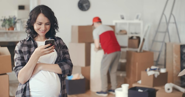 Portret szczęśliwej kobiety w ciąży używa telefonu komórkowego w nowym mieszkaniu