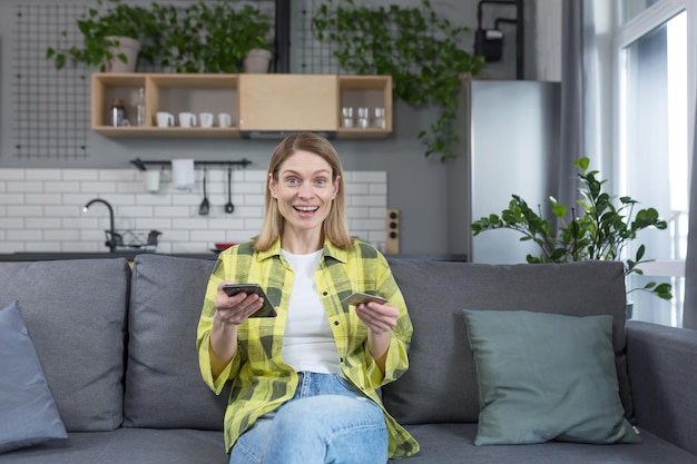 Zdjęcie portret szczęśliwej kobiety w domu gospodyni siedząca na kanapie szczęśliwa i patrząca na kamerę uśmiechnięta trzymająca telefon i bankową kartę kredytową na zakupy online